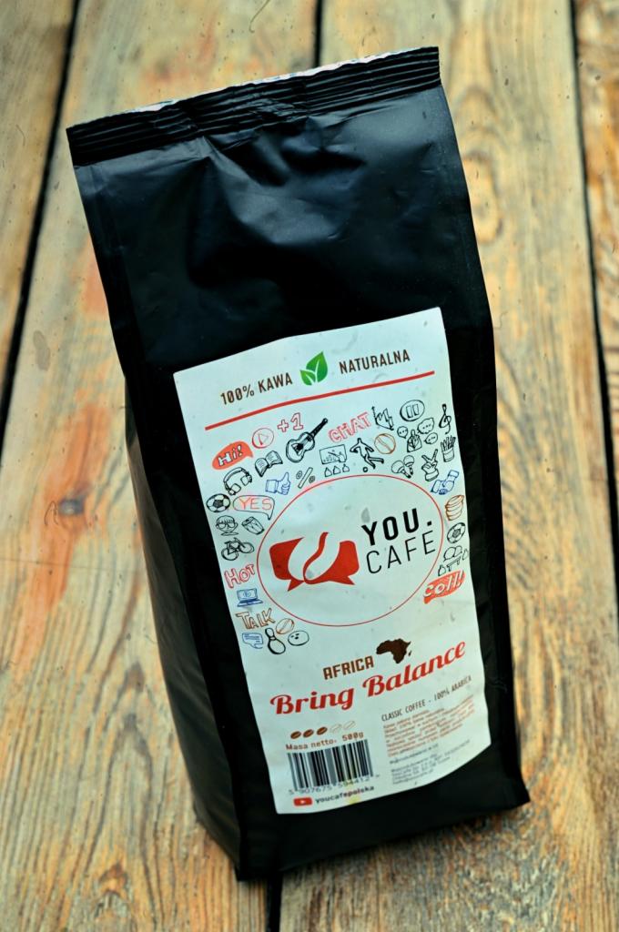 Czarna paczka kawy ziarnistej You.Cafe Africa Bring Balance, leżąca na drewnianym blacie z widocznym logo i informacjami o produkcie.