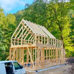 Konstrukcja szkieletowa domu w otoczeniu drzew, z białym vanem zaparkowanym z przodu, widok na fundament i częściowo ukończony szkielet dachu.