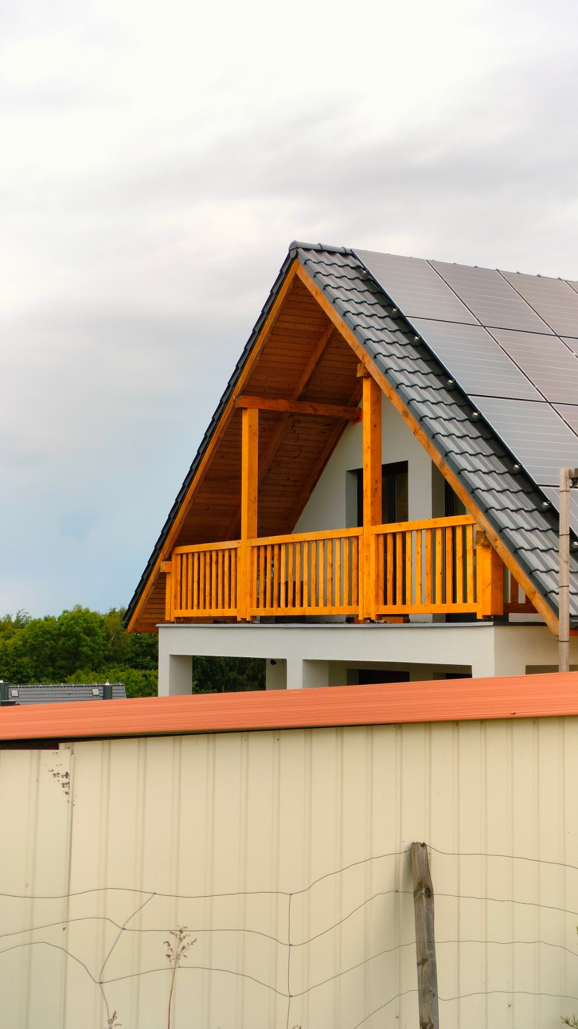 Dwukondygnacyjny dom z drewnianym balkonem i panelami słonecznymi na dachu, widok zza ogrodzenia z siatką.
