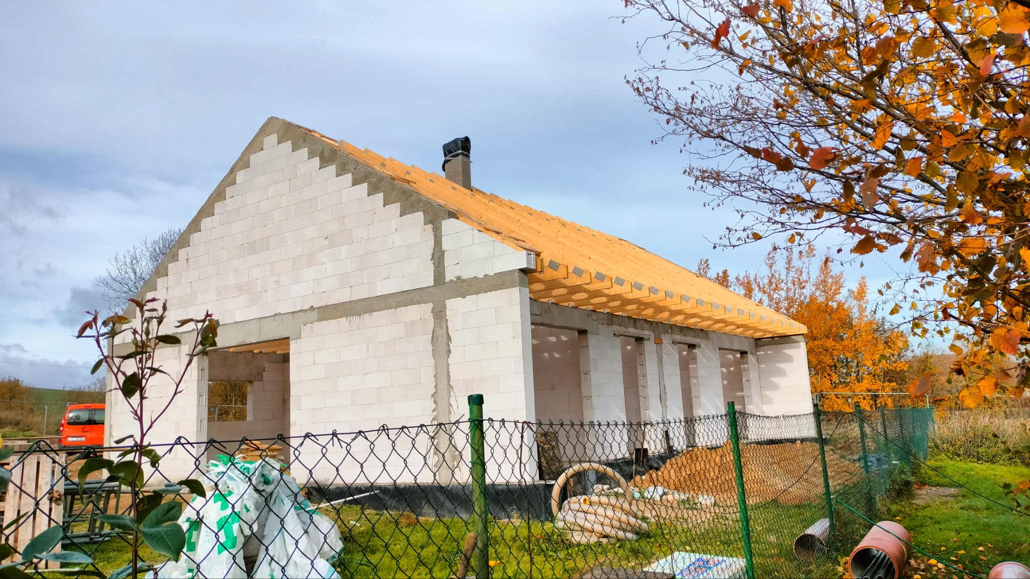 Surowy stan domu z białego betonu komórkowego, z drewnianą konstrukcją dachu w trakcie budowy, widoczne zza metalowego ogrodzenia i jesiennego drzewa.