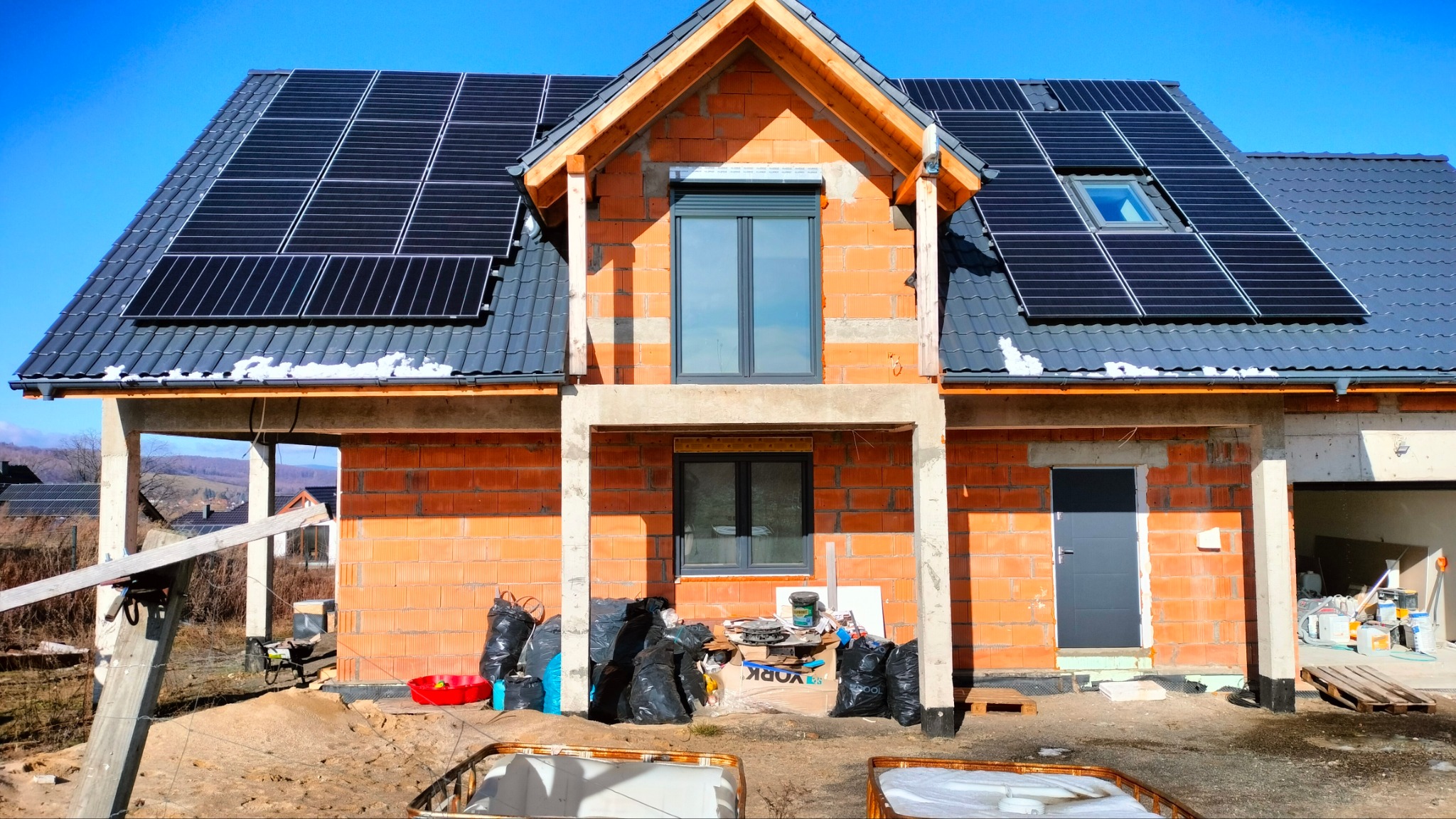 Niedokończony dom z czerwonej cegły z zamontowanymi panelami słonecznymi na dachu i oknem dachowym; widoczne rusztowanie, worki z odpadami budowlanymi i narzędzia.