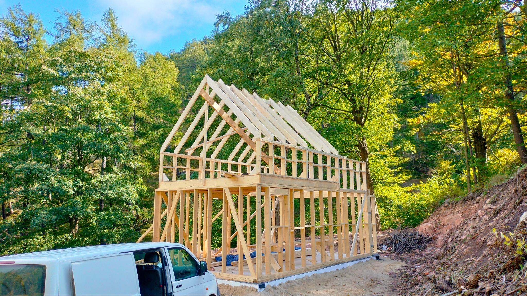 Konstrukcja szkieletowa domu w otoczeniu drzew, z białym vanem zaparkowanym z przodu, widok na fundament i częściowo ukończony szkielet dachu.