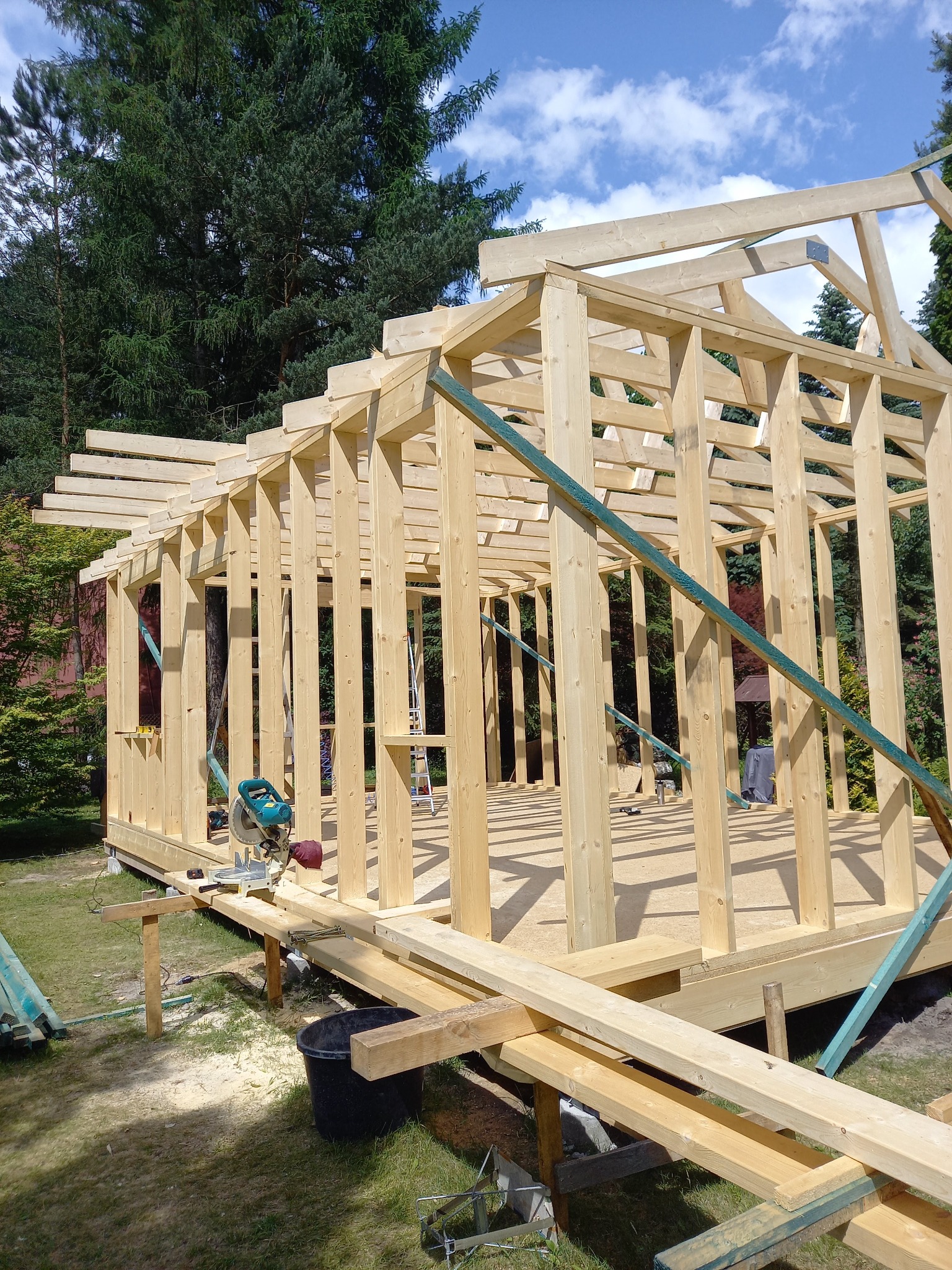 Surowa drewniana konstrukcja domu w trakcie budowy, widoczne elementy szkieletu, narzędzia i piła stołowa na zewnątrz, w tle drzewa i niebo.
