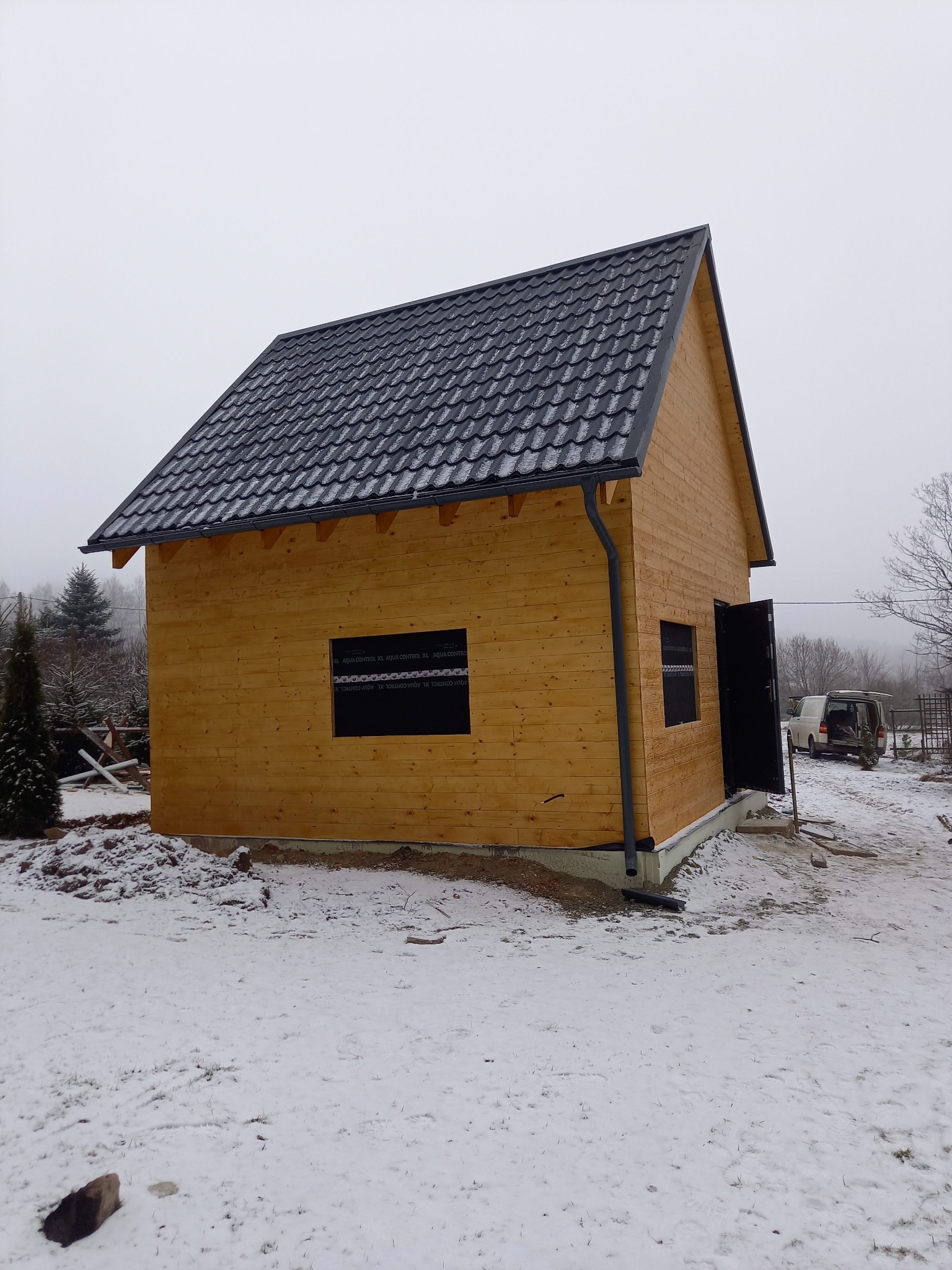 Nowo wybudowany, drewniany domek gospodarczy z czarną, metalową dachówką, częściowo ośnieżony, z otwartymi drzwiami wejściowymi i folią okienną, na tle zimowego krajobrazu.