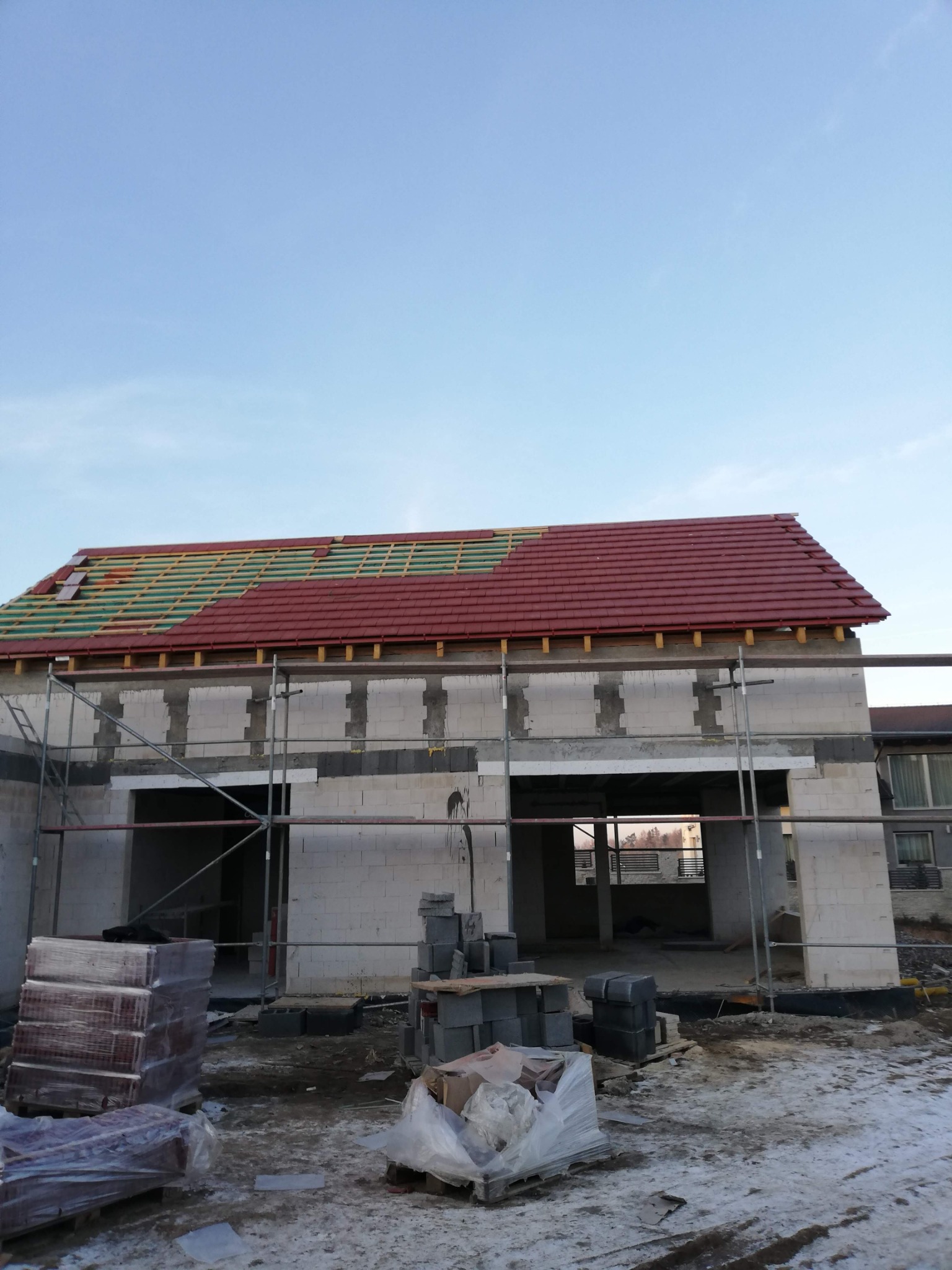 Budowa domu z pustaków: częściowo ułożona czerwona dachówka na drewnianej konstrukcji, widoczne rusztowania i materiały budowlane na placu budowy.