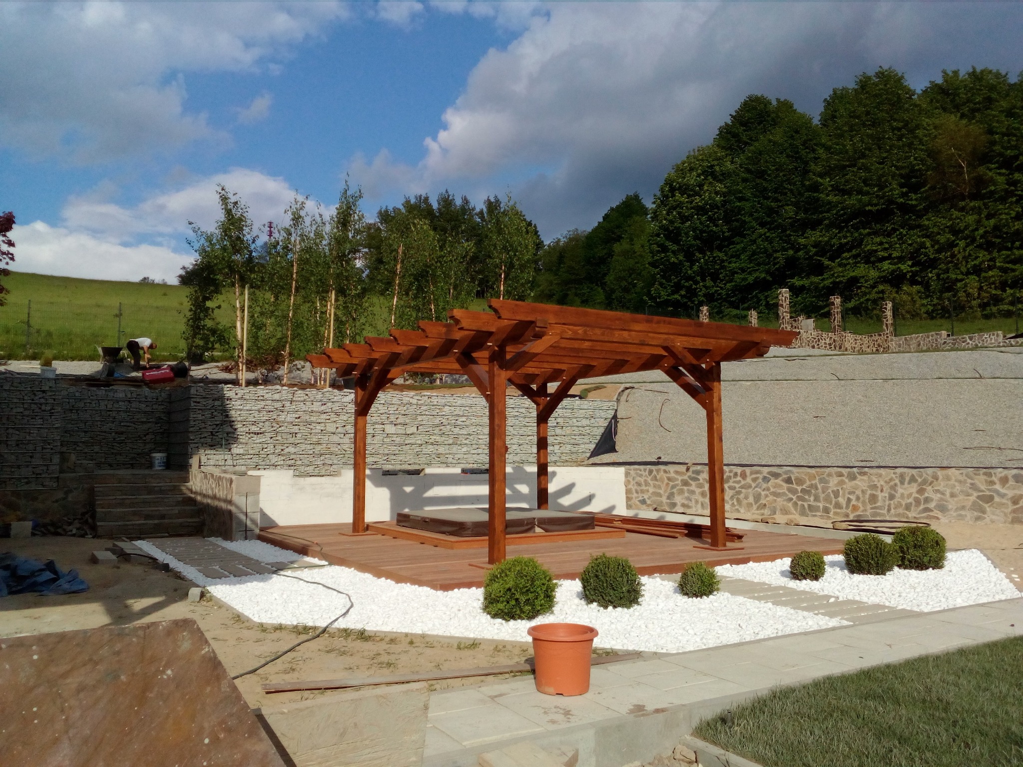 Drewniana pergola nad jacuzzi na tarasie, otoczona białym żwirem i niskimi krzewami, w tle mur z kamienia naturalnego i zielone drzewa.