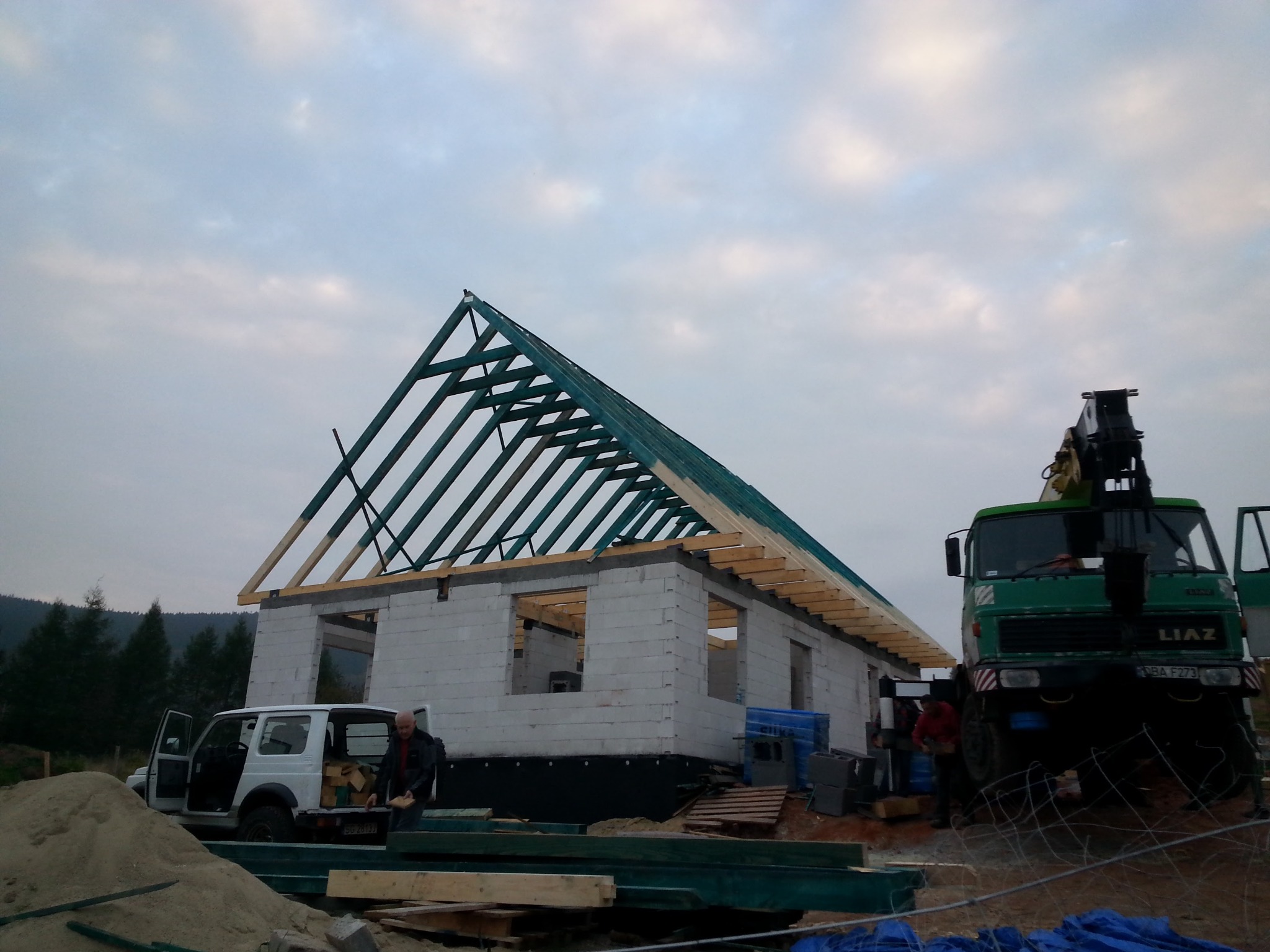 Wznoszenie domu z betonu komórkowego: ściany, więźba dachowa z zieloną impregnacją, dźwig budowlany i Suzuki Jimny na placu budowy.