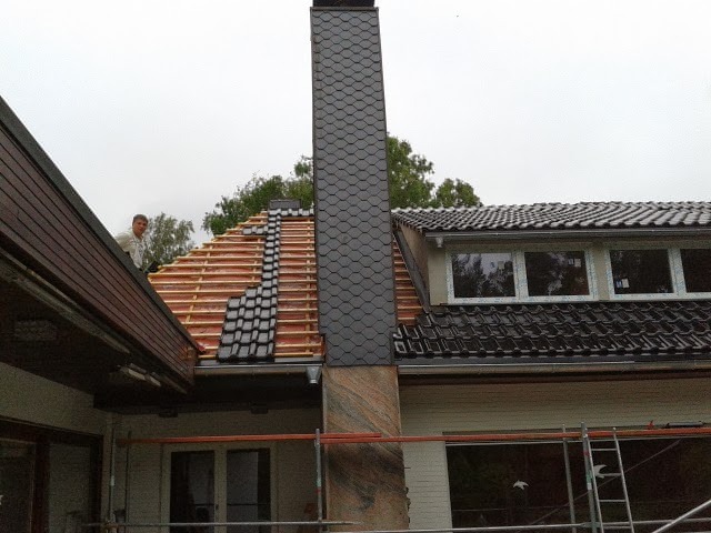 Układanie dachówki na nowym dachu domu jednorodzinnego, widoczny komin pokryty łupkiem i pracownik na dachu, częściowo ułożona dachówka oraz konstrukcja drewniana.