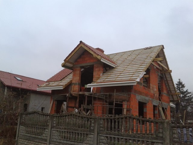 Surowy, dwukondygnacyjny dom z czerwonej cegły w trakcie budowy, widoczne drewniane elementy konstrukcji dachu oraz częściowo ułożona dachówka, szara betonowa podmurówka w tle.