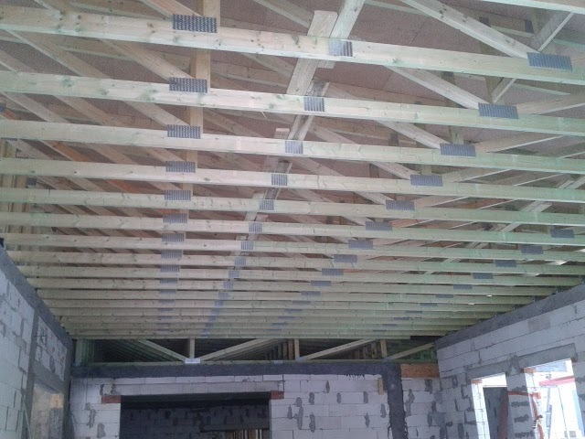 Wnętrze domu w trakcie budowy, widoczne drewniane belki stropowe połączone metalowymi płytkami kolczastymi, ściany z bloczków betonowych.