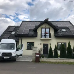 Dom jednorodzinny z zamontowanymi panelami fotowoltaicznymi na dachu, biały bus z otwartymi drzwiami na podjeździe, pochmurne niebo w tle.