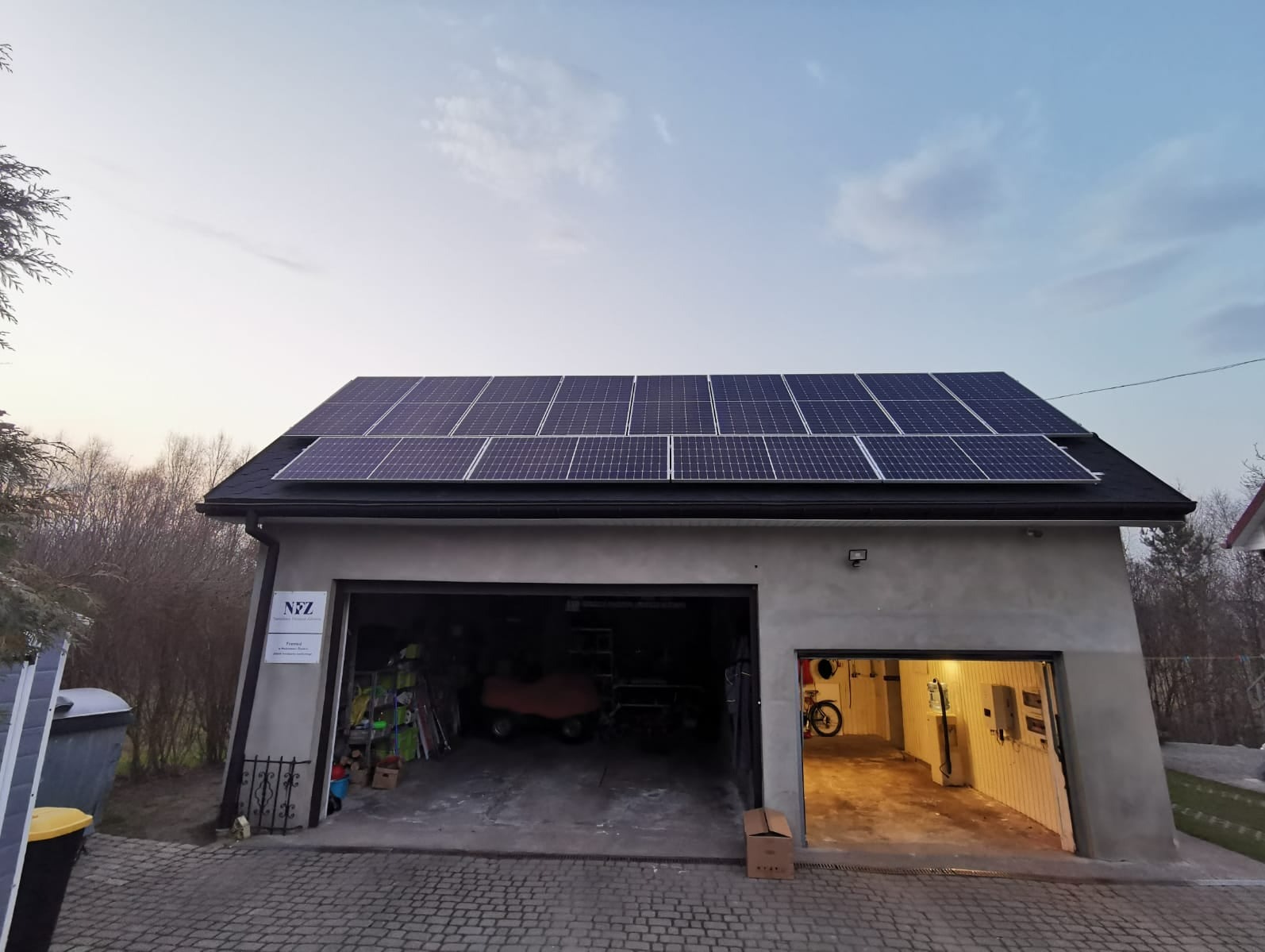 Panele słoneczne na dachu garażu z dwoma otwartymi bramami, widoczny sprzęt wewnątrz, tabliczka z logo firmy na ścianie.