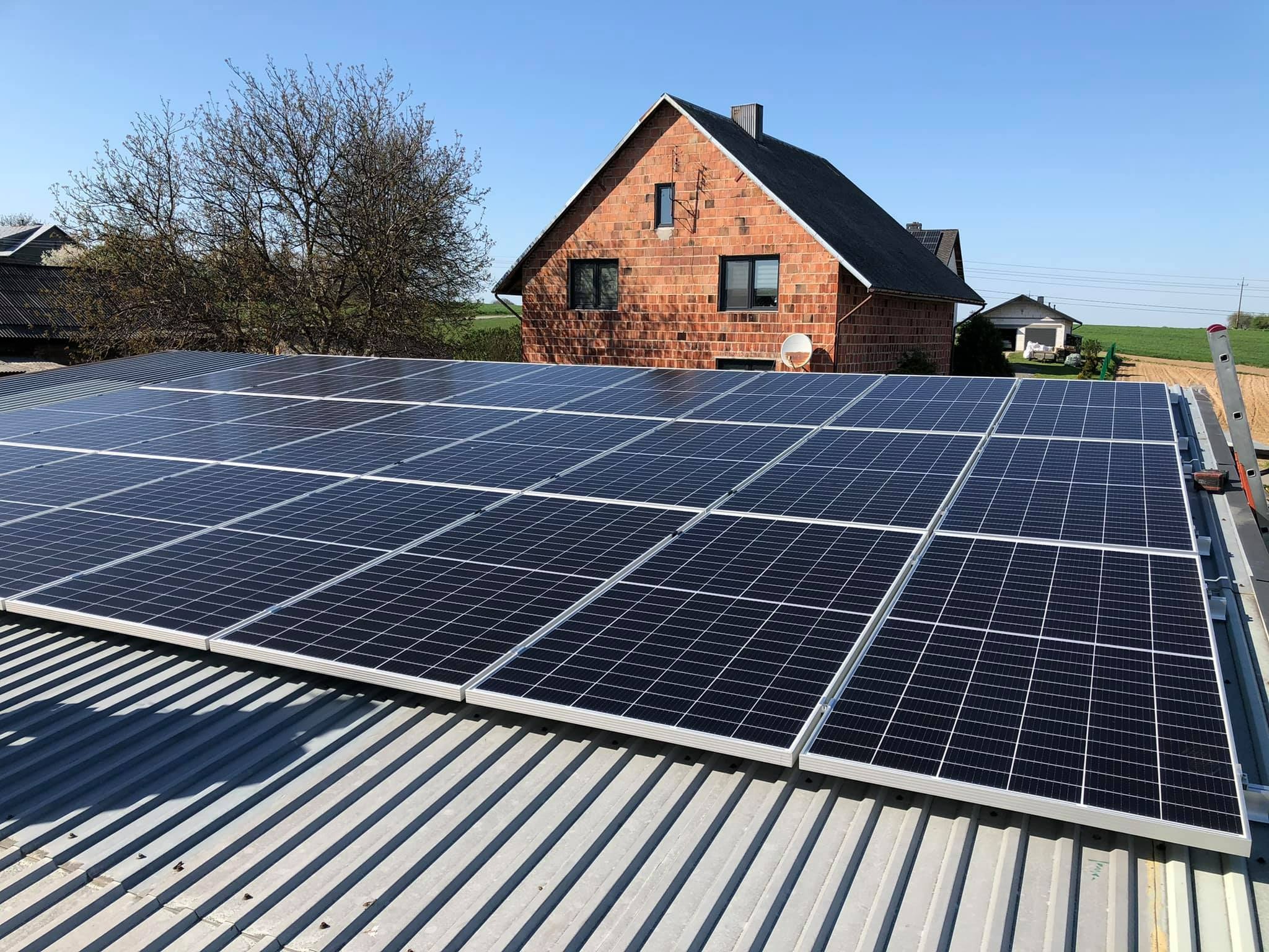 Instalacja paneli słonecznych na falistym blaszanym dachu domu z czerwonej cegły w słoneczny dzień.