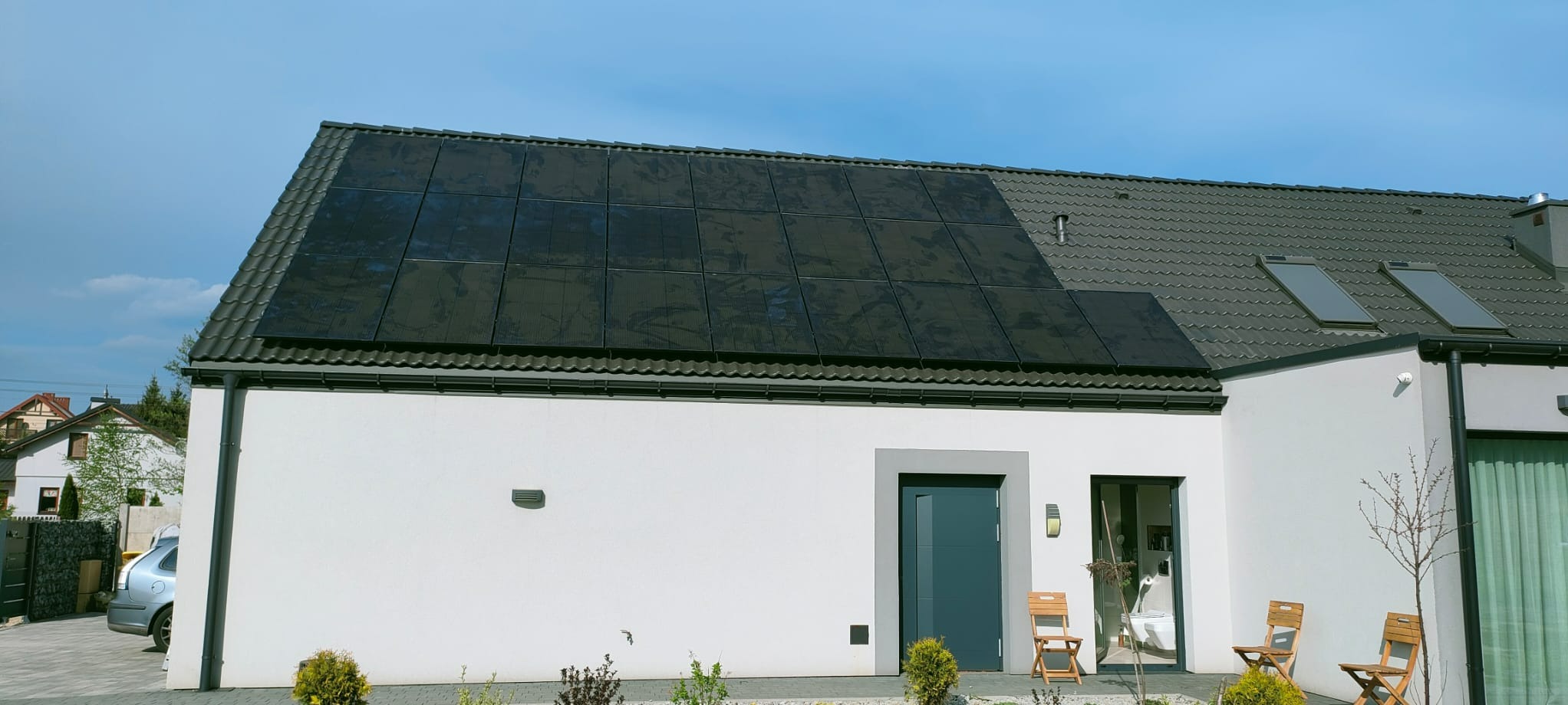 Dom jednorodzinny z panelami słonecznymi na dachu pokrytym szarą dachówką, widoczne okna dachowe, biała elewacja, szare drzwi wejściowe i okno, drewniane krzesła ogrodowe przed wejściem.