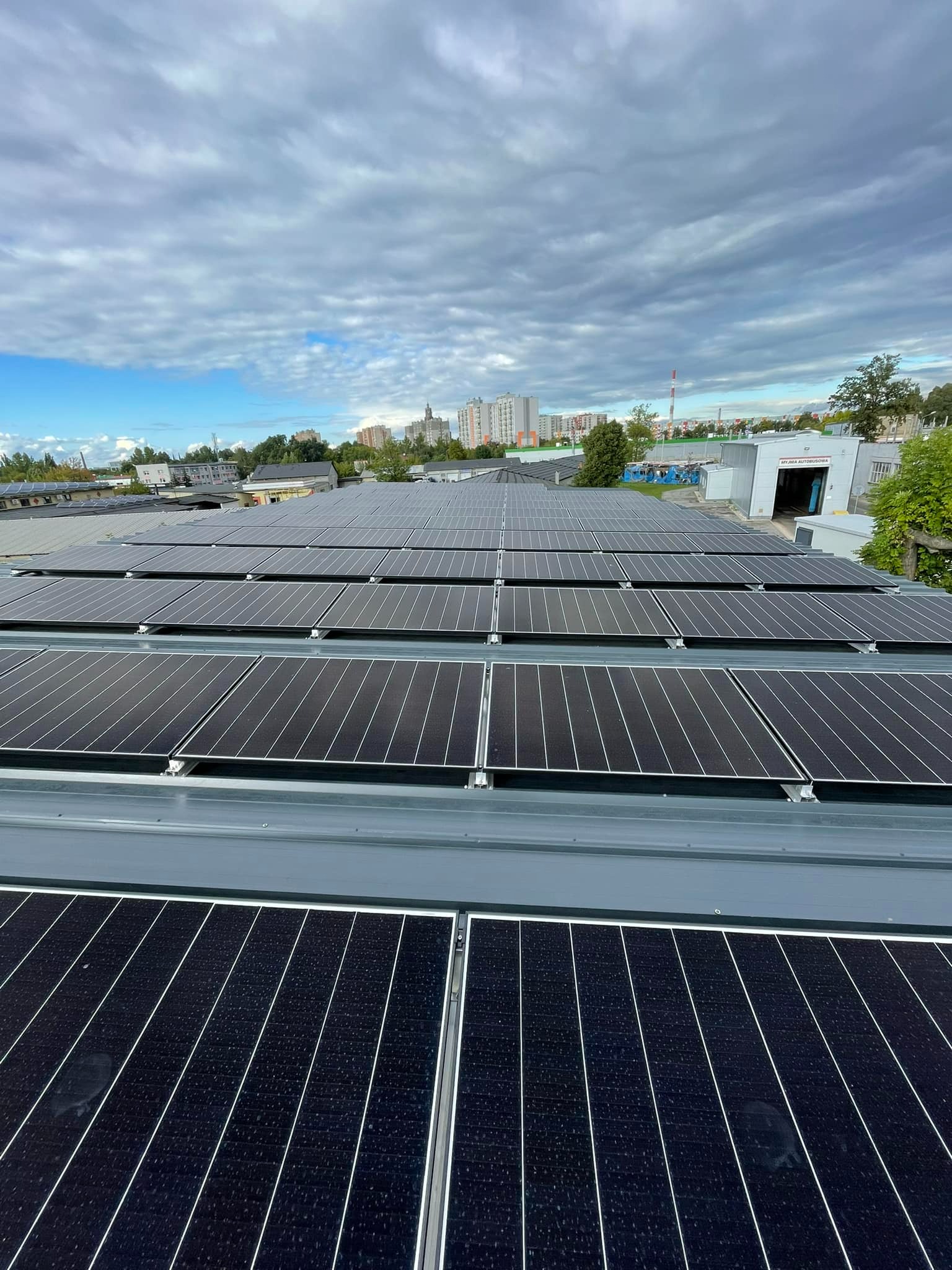 Rząd paneli słonecznych na dachu budynku przemysłowego, widok z góry na instalację i panoramę miasta w tle pod pochmurnym niebem.