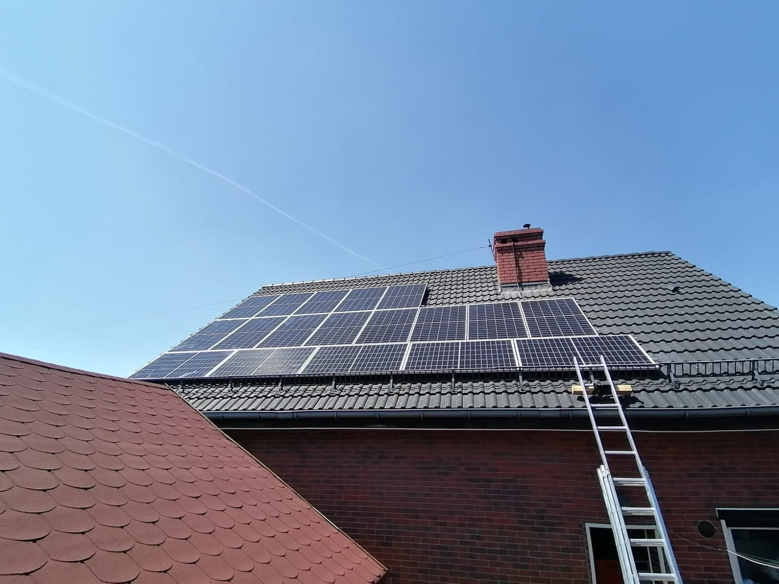 Panele słoneczne zamontowane na dachu domu z cegły, drabina oparta o dach, błękitne niebo w tle.