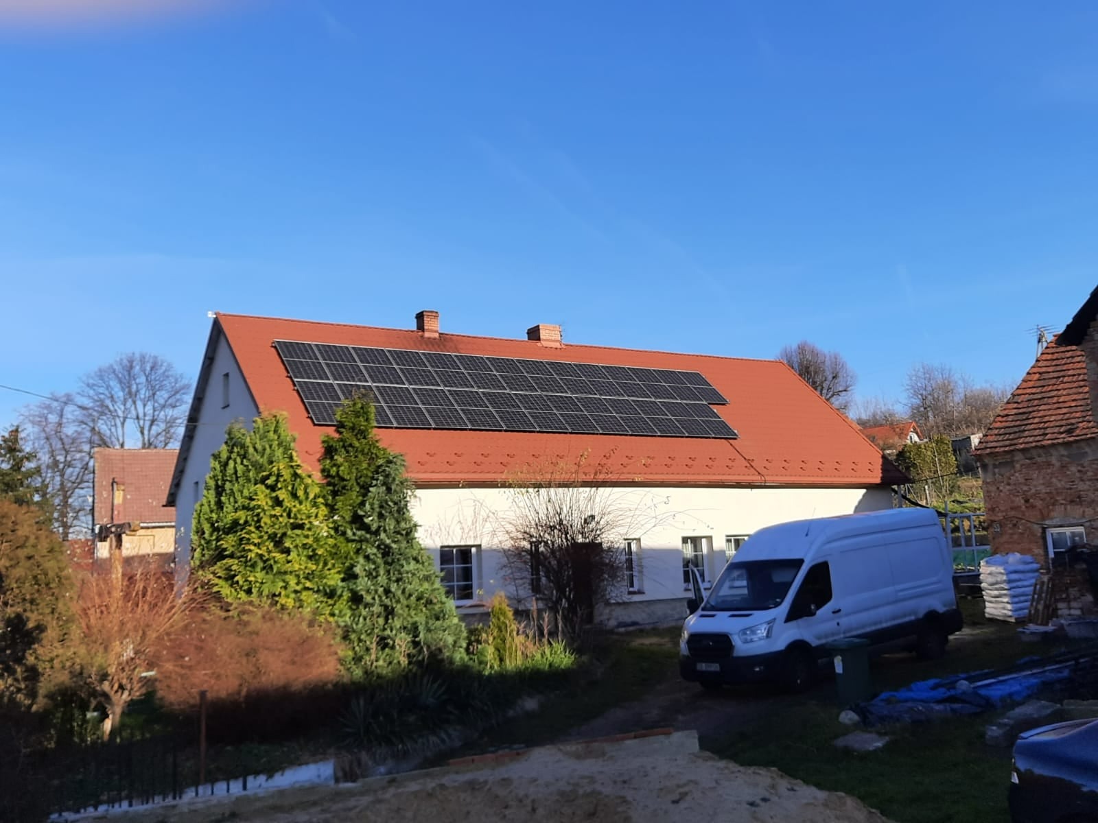 Dom jednorodzinny z czerwoną dachówką, na której zamontowano panele słoneczne. Biały samochód dostawczy parkuje obok budynku. Widoczne worki z materiałami budowlanymi.