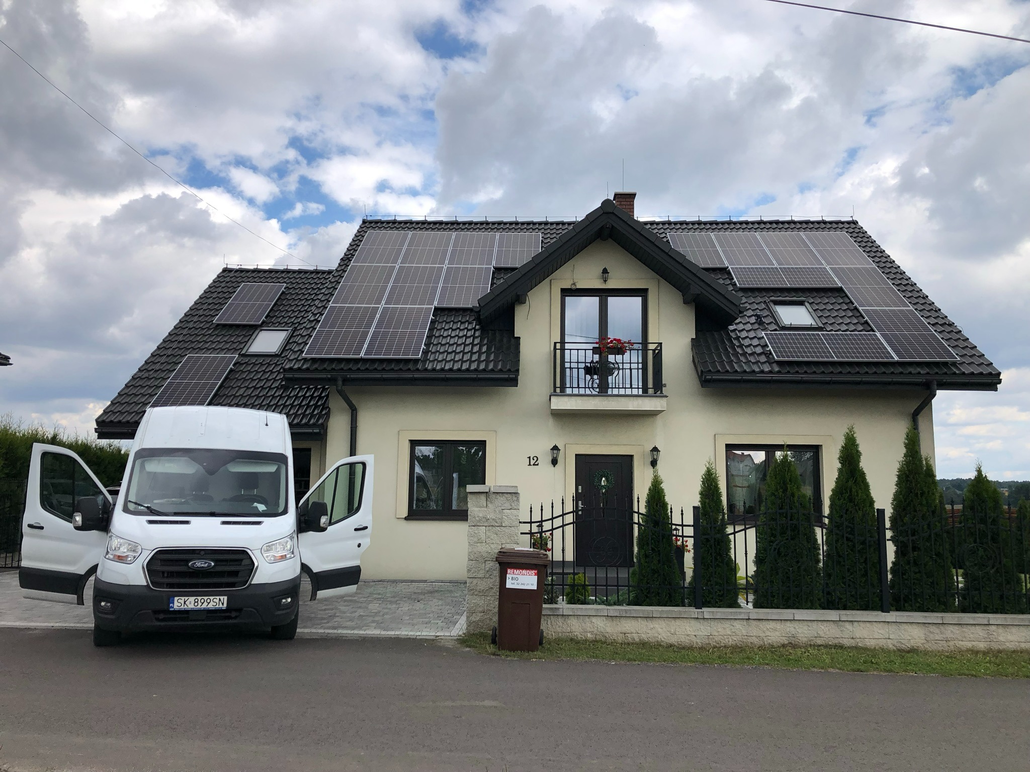 Dom jednorodzinny z zamontowanymi panelami fotowoltaicznymi na dachu, biały bus z otwartymi drzwiami na podjeździe, pochmurne niebo w tle.