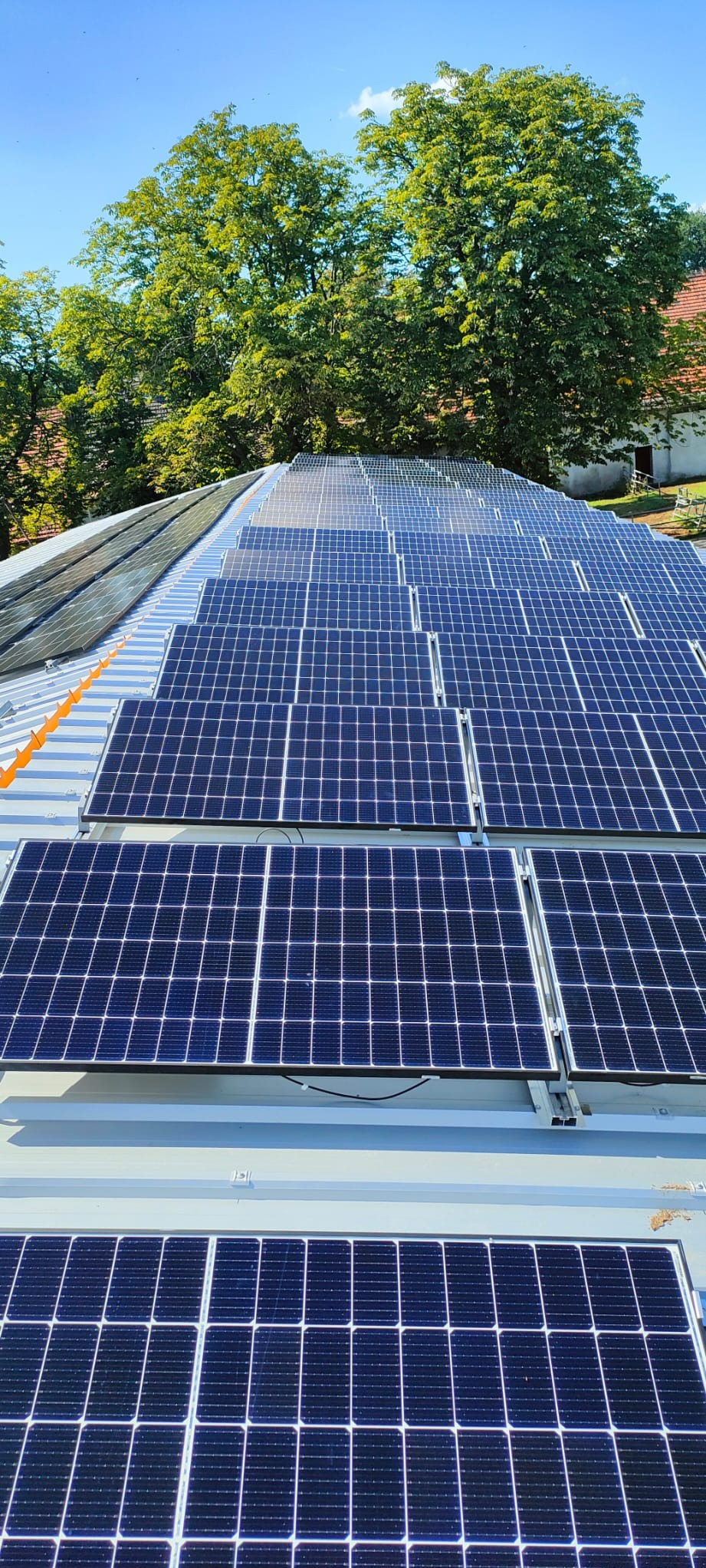 Rząd paneli słonecznych na metalowym dachu, widoczne mocowania i drzewa w tle.