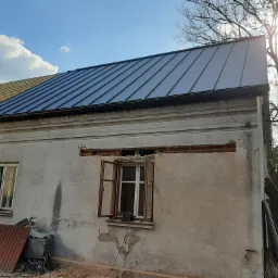 Fragment domu z częściowo wymienionym dachem z blachy trapezowej w kolorze grafitowym; widoczne uszkodzenia elewacji, otwarte okno z narzędziami na parapecie oraz drzewo bez liści.