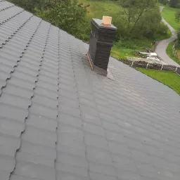 Szary dach z blachy trapezowej z kominem z cegły klinkierowej na tle zieleni i zabudowań.