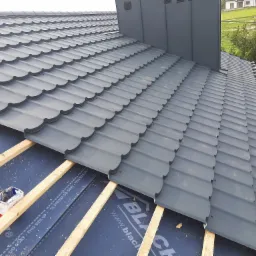 Nowy, szary dach z blachodachówki, częściowo ułożony na łatach, widoczna membrana dachowa z napisami i otwarty pojemnik z elementami mocującymi, komin w kolorze dachu, w tle dom i zieleń.