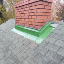 Ceglany komin z nową, zieloną obróbką blacharską na dachu pokrytym gontem bitumicznym, widok z góry.