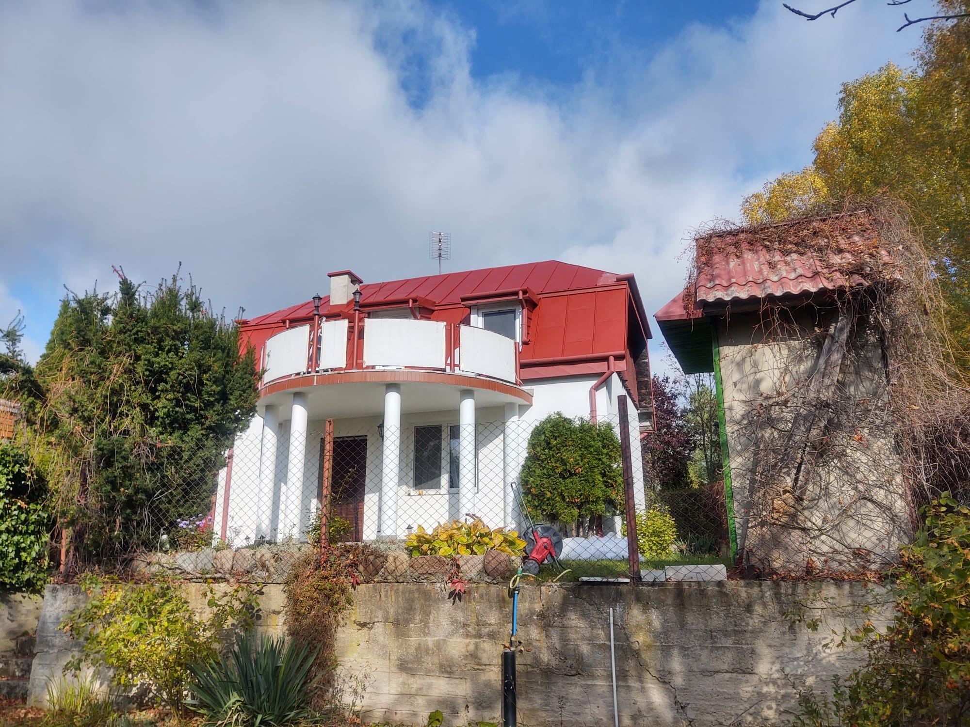 Dwupiętrowy dom z czerwonym dachem blacharskim, białymi ścianami i zaokrąglonym balkonem, widoczny z dołu przez ogrodzenie z siatki.