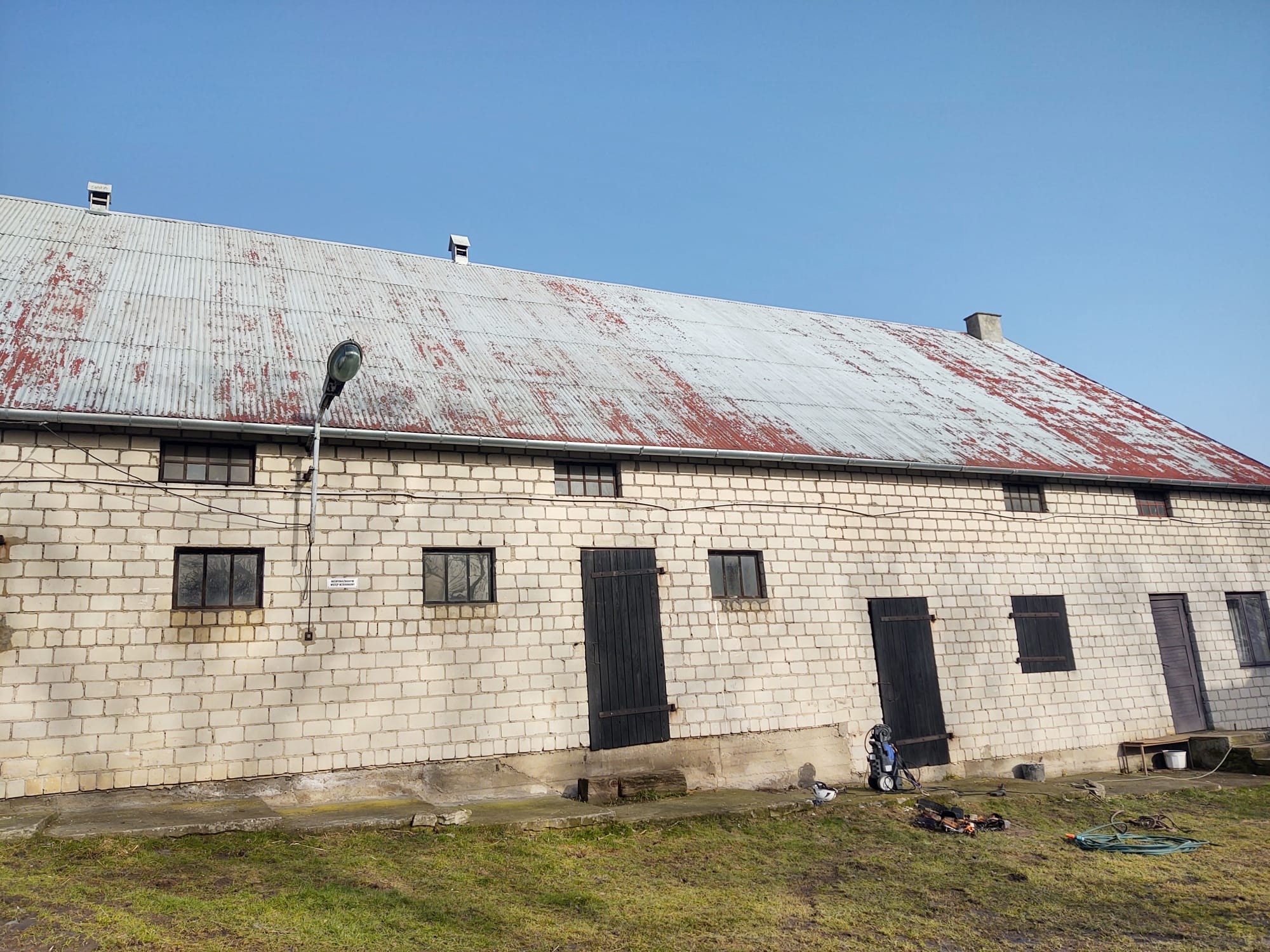 Budynek gospodarczy z blachy falistej na dachu, widoczne ślady korozji, ściany z białej cegły, czarne okna i drzwi, na trawie leży wąż ogrodowy i myjka ciśnieniowa.