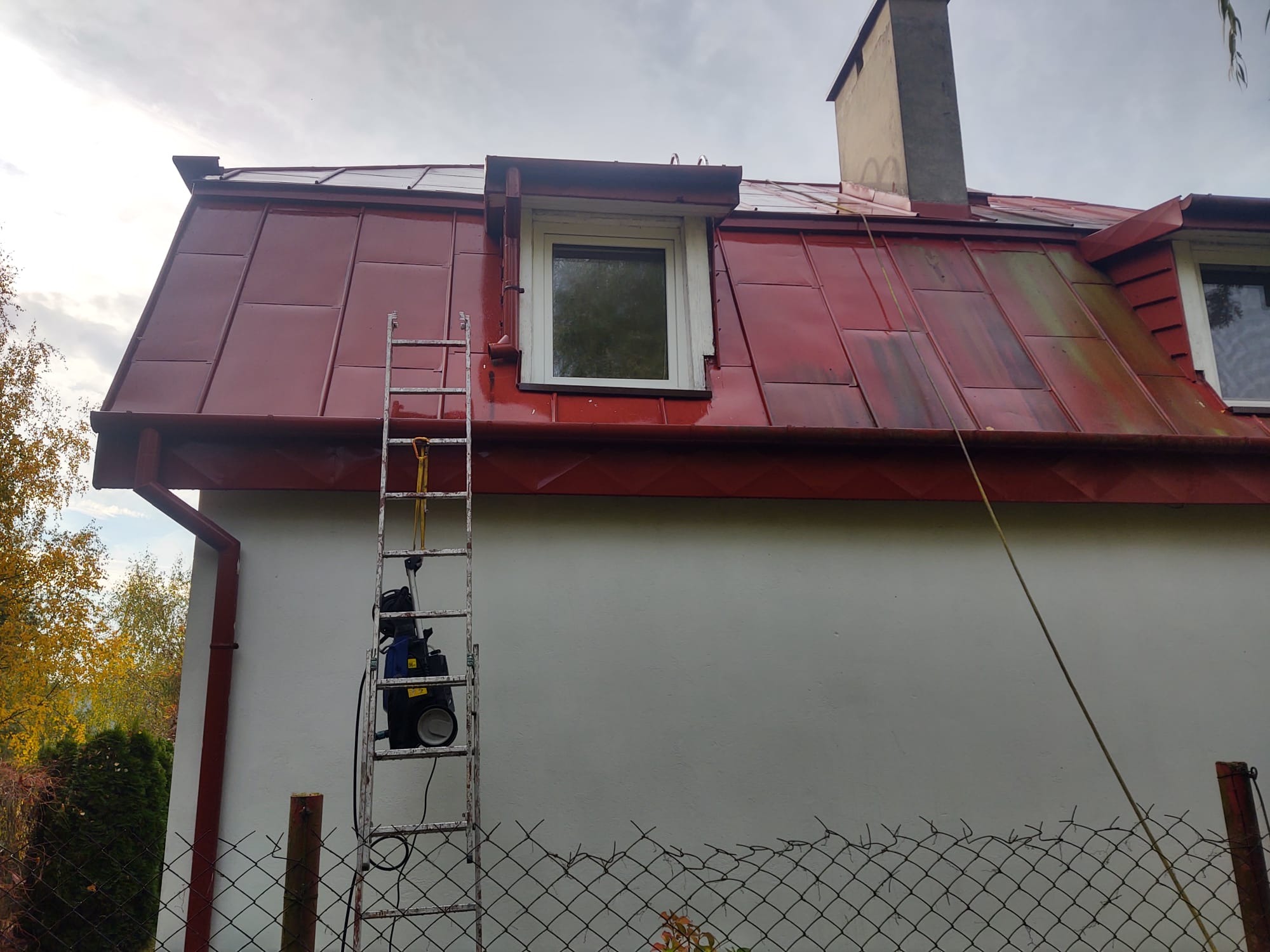 Czerwony dach z blachy trapezowej po czyszczeniu, widoczna drabina oparta o ścianę budynku z myjką ciśnieniową, fragment rynny i komina.