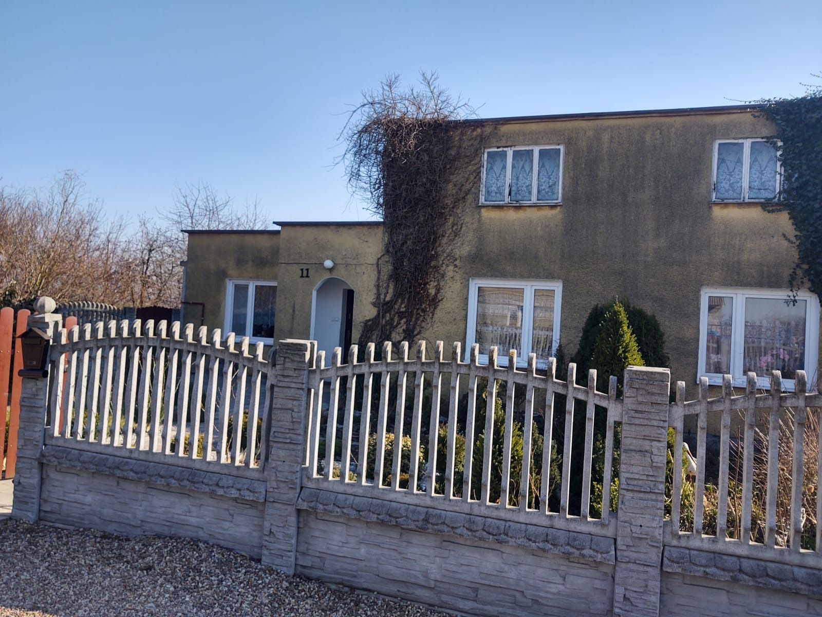 Dwukondygnacyjny dom z szarą elewacją, porośnięty pnączami przy wejściu i w prawym górnym rogu, z betonowym ogrodzeniem i numerem 11 nad wejściem.