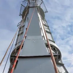 Wysoka, metalowa konstrukcja pomalowana na szaro, widziana z dołu, z linami zwisającymi z góry i niebieskim niebem w tle.