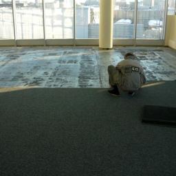 Budex Group - Pracownik w kombinezonie montuje kwadratowe płytki wykładzinowe w jasnym pomieszczeniu z dużymi oknami, widoczne przygotowane podłoże.