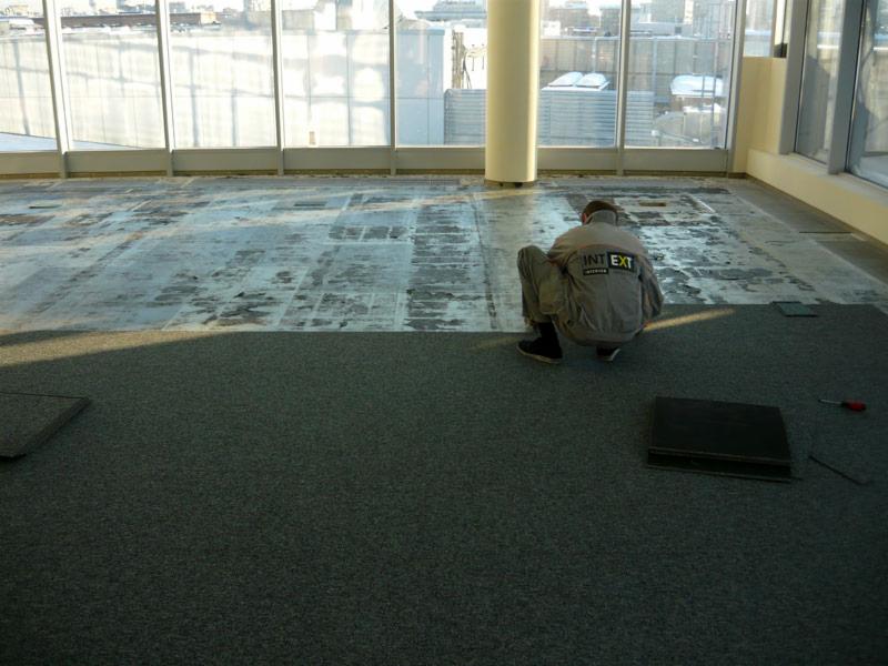 Pracownik w kombinezonie montuje kwadratowe płytki wykładzinowe w jasnym pomieszczeniu z dużymi oknami, widoczne przygotowane podłoże.