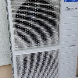 Biała zewnętrzna jednostka klimatyzacji Panasonic Inverter zamontowana przy ścianie budynku, widoczne otwory wentylacyjne i logo producenta.