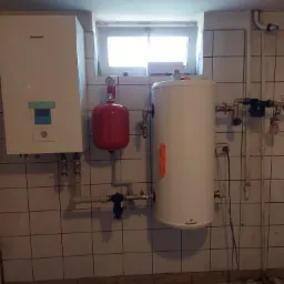 Instalacja z białym zbiornikiem akumulacyjnym, czerwonym naczyniem przeponowym i jednostką zewnętrzną w pomieszczeniu z kafelkami w Ostrowi Mazowieckiej. Na podłodze widoczne narzędzia i elementy...