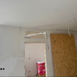 Climca-M Kamil Żabik - Wnętrze w trakcie budowy z widocznymi przewodami wentylacyjnymi lub klimatyzacyjnymi nad otworem drzwiowym, wiszącymi kablami z sufitu i płytą OSB.