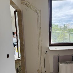 Climca-M Kamil Żabik - Wnętrze w trakcie remontu z bruzdami w ścianie, w których ułożono przewody instalacyjne, częściowo zabezpieczone pianką. Widoczne okno z widokiem na miasto i grzejnik.