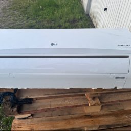 Climca-M Kamil Żabik - Biała jednostka wewnętrzna klimatyzatora LG Inverter V, z widocznymi trybami pracy, leżąca na drewnianych paletach na zewnątrz, z trawą i metalowym ogrodzeniem w tle.