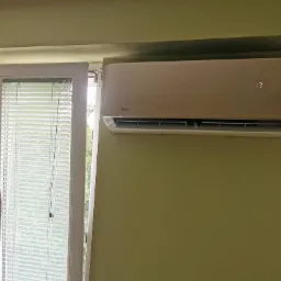 Beżowy klimatyzator marki Vivax zamontowany na zielonej ścianie w pomieszczeniu z oknem z żaluzjami, widoczny wyświetlacz z temperaturą 17 stopni Celsjusza.