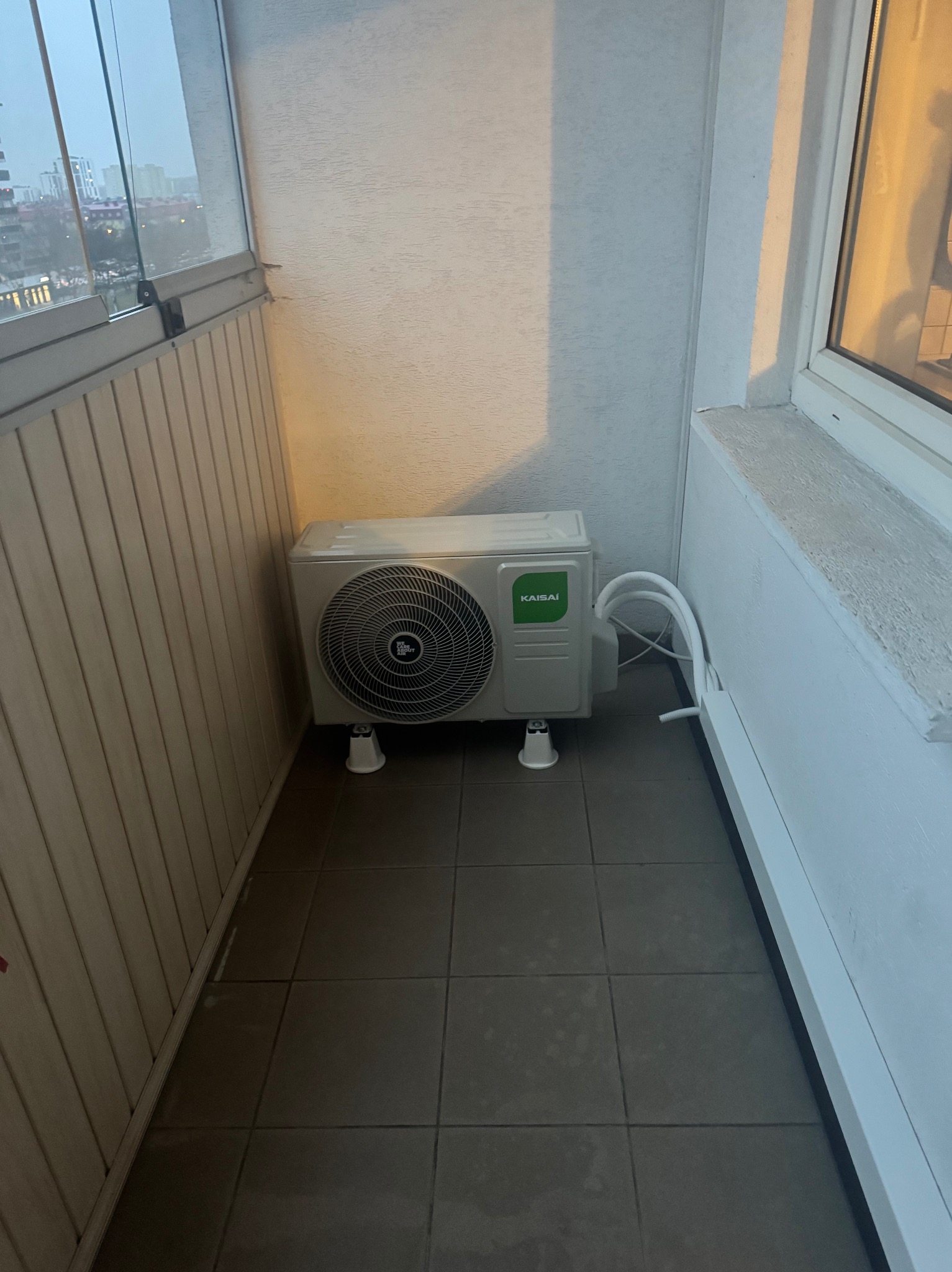 Kompaktowy klimatyzator Kaisai na balkonie z szarą podłogą z płytek. Widoczne białe rury i elementy montażowe. Za oknem miejski krajobraz w tle.