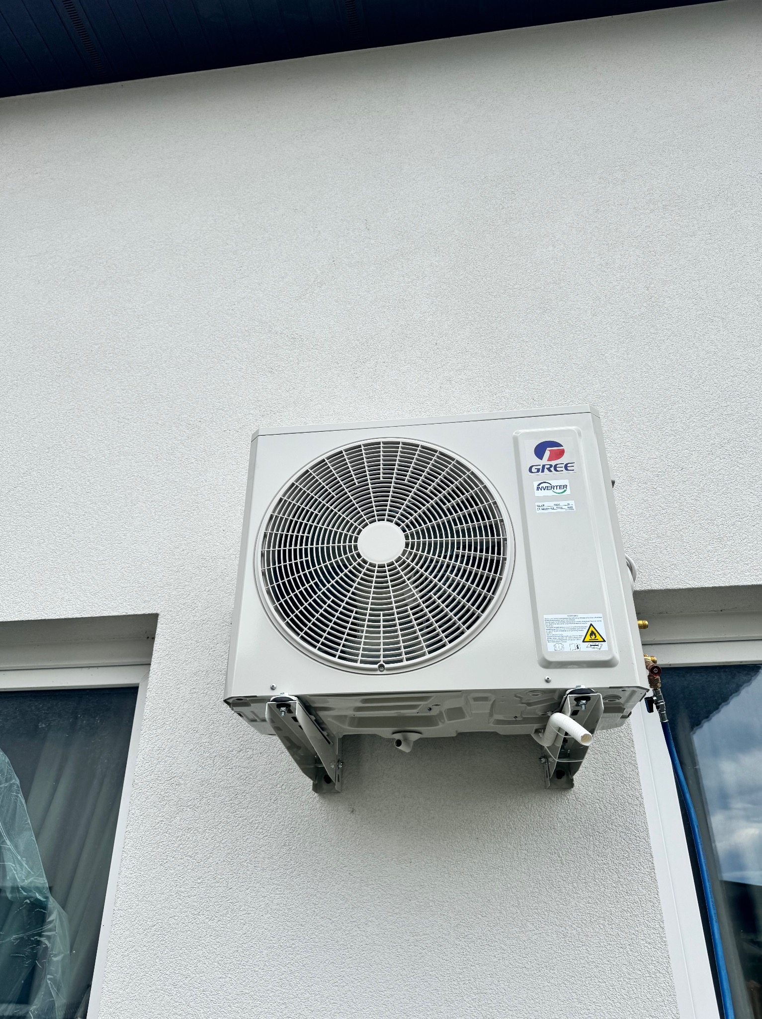 Zewnętrzna jednostka klimatyzatora GREE Inverter zamontowana na białej, fakturowanej ścianie budynku, obok okna z folią ochronną.