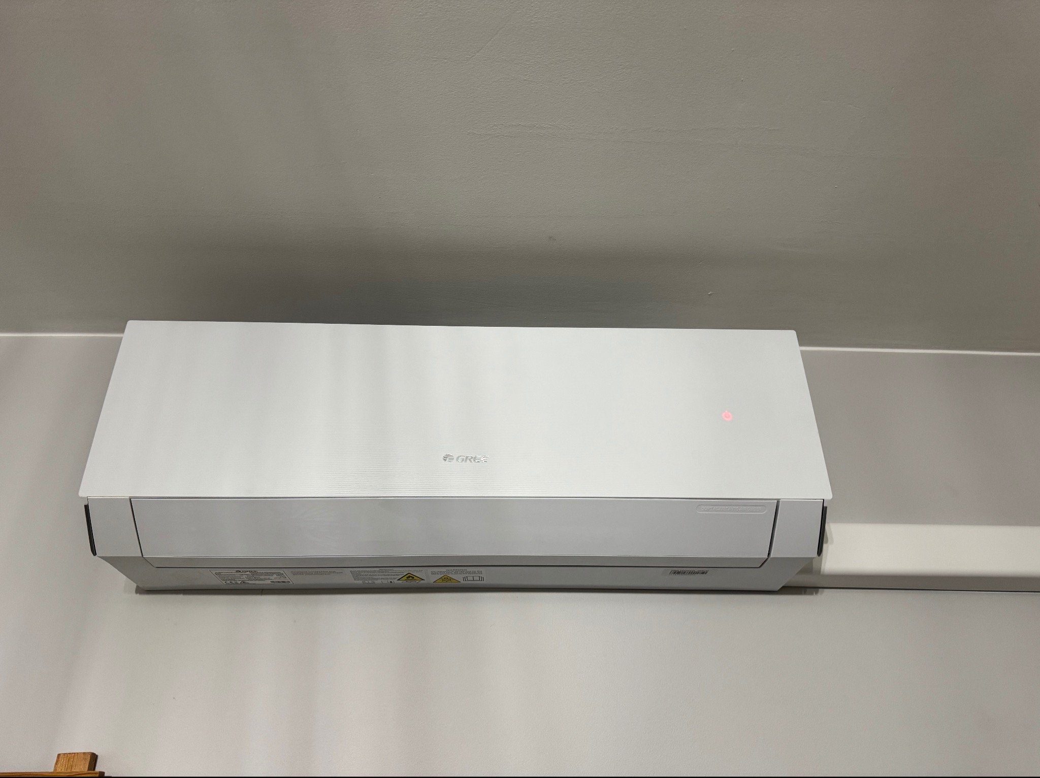 Biała jednostka klimatyzacyjna GREE zamontowana na jasnoszarej ścianie, z widocznym czerwonym wskaźnikiem i białym korytem instalacyjnym po prawej.