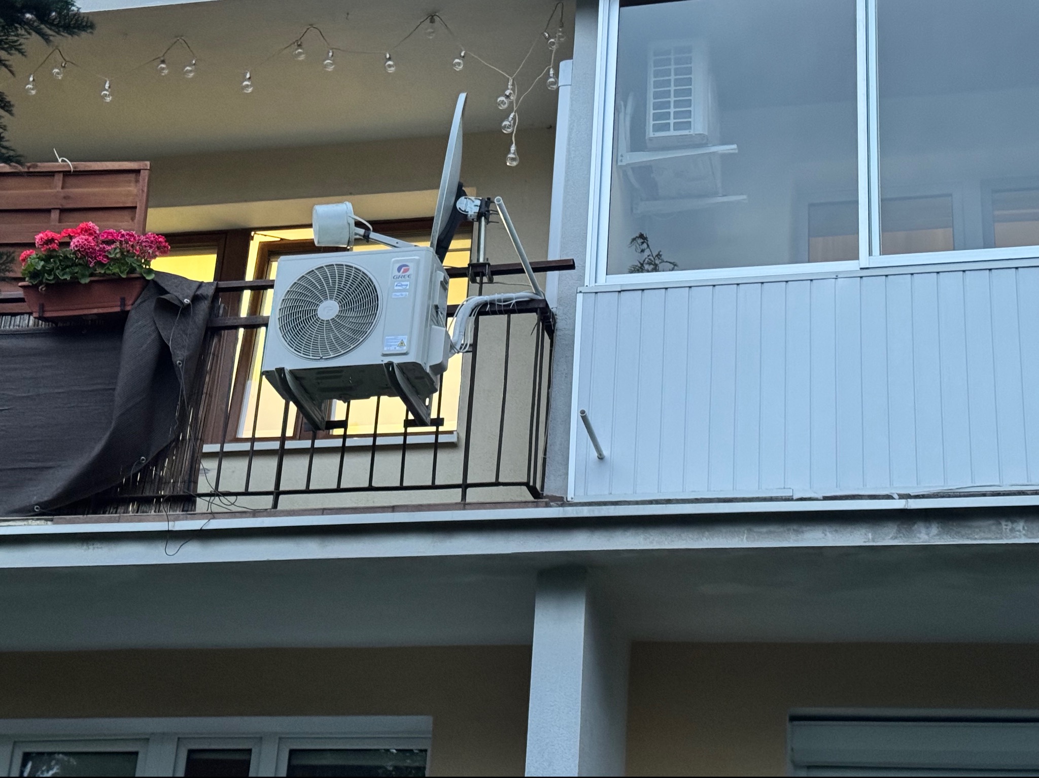 Zewnętrzna jednostka klimatyzatora marki Gree zamontowana na balustradzie balkonu, obok anteny satelitarnej, z doniczką różowych kwiatów i girlandą światełek w tle.