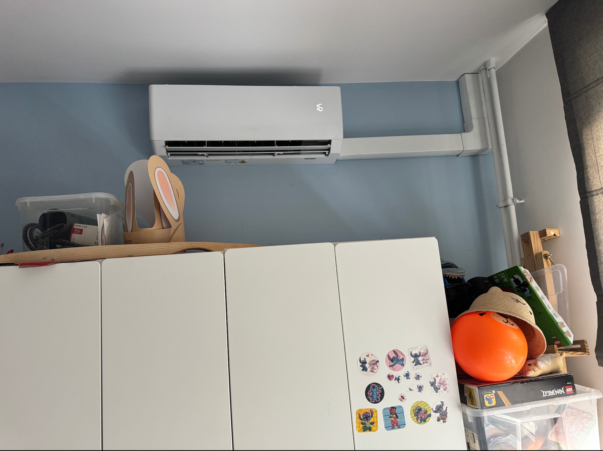 Biała klimatyzacja ścienna z wyświetlaczem '16' zamontowana na jasnoniebieskiej ścianie, z białym korytem instalacyjnym i rurą, nad szafkami z naklejkami.