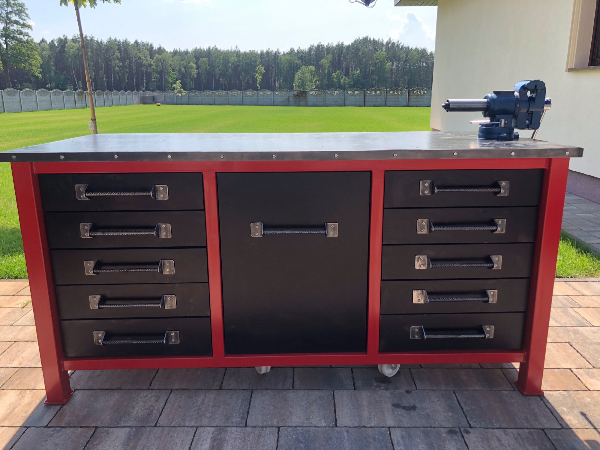 Czerwono-czarny warsztatowy stół z metalowym blatem i imadłem, z widocznymi uchwytami szuflad wykonanymi ze skręconego metalu, na tle trawnika i drzew.