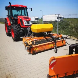 Czerwony ciągnik Zetor z pomarańczowym osprzętem do pielęgnacji zieleni, w tle białe ciężarówki i błękitne niebo.