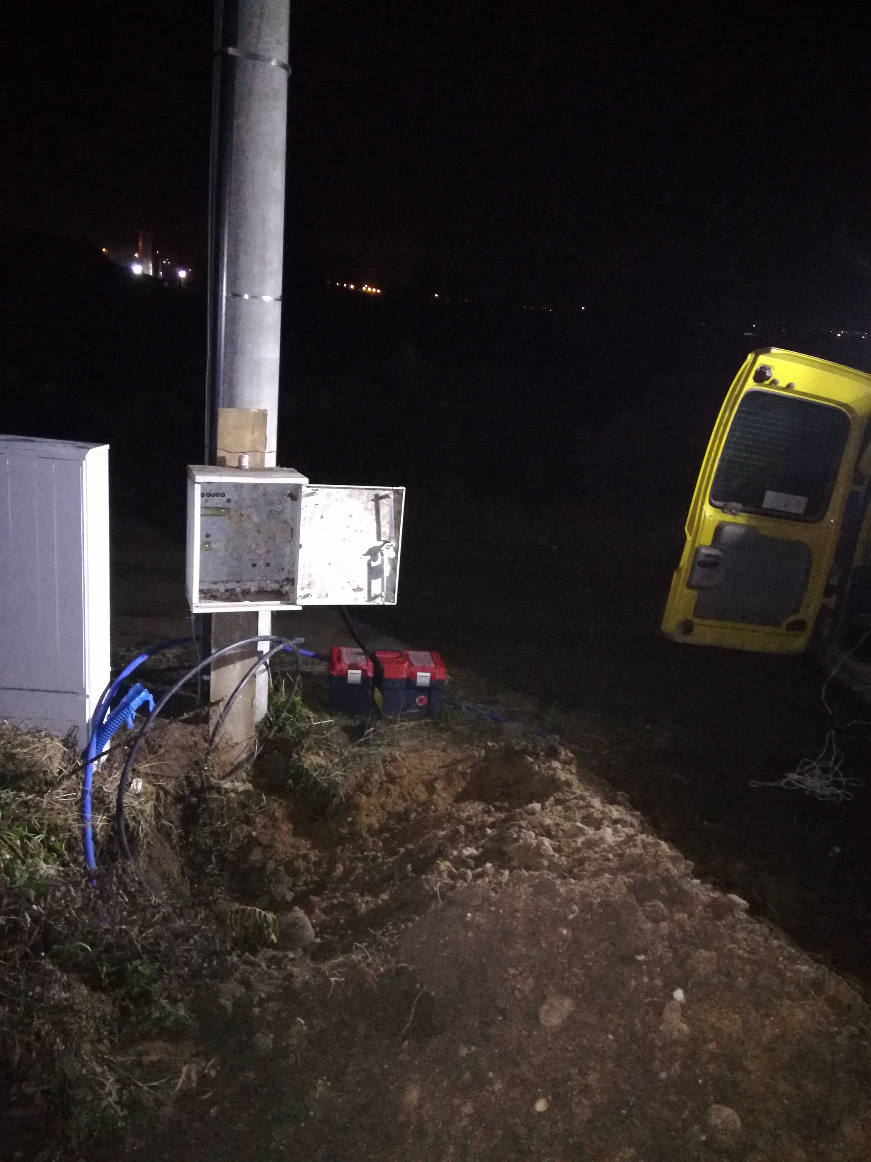 Instalacja elektryczna zewnętrzna w trakcie realizacji: skrzynka elektryczna z otwartymi drzwiczkami, kable w peszlach, skrzynki narzędziowe i żółty samochód dostawczy w tle, widok nocny.