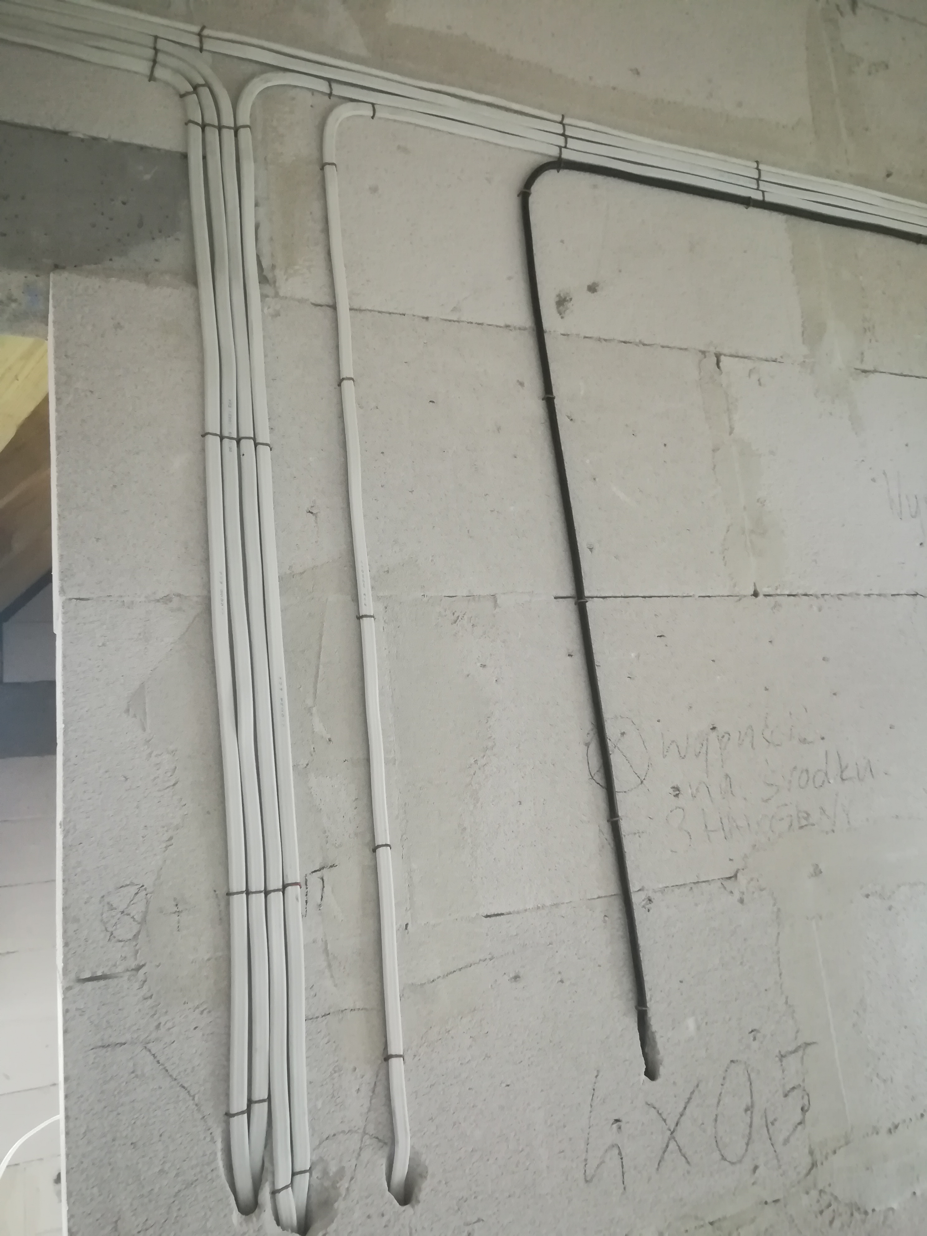 Białe i czarne przewody elektryczne prowadzone po szarej ścianie z betonu komórkowego, zabezpieczone czarnymi opaskami zaciskowymi.