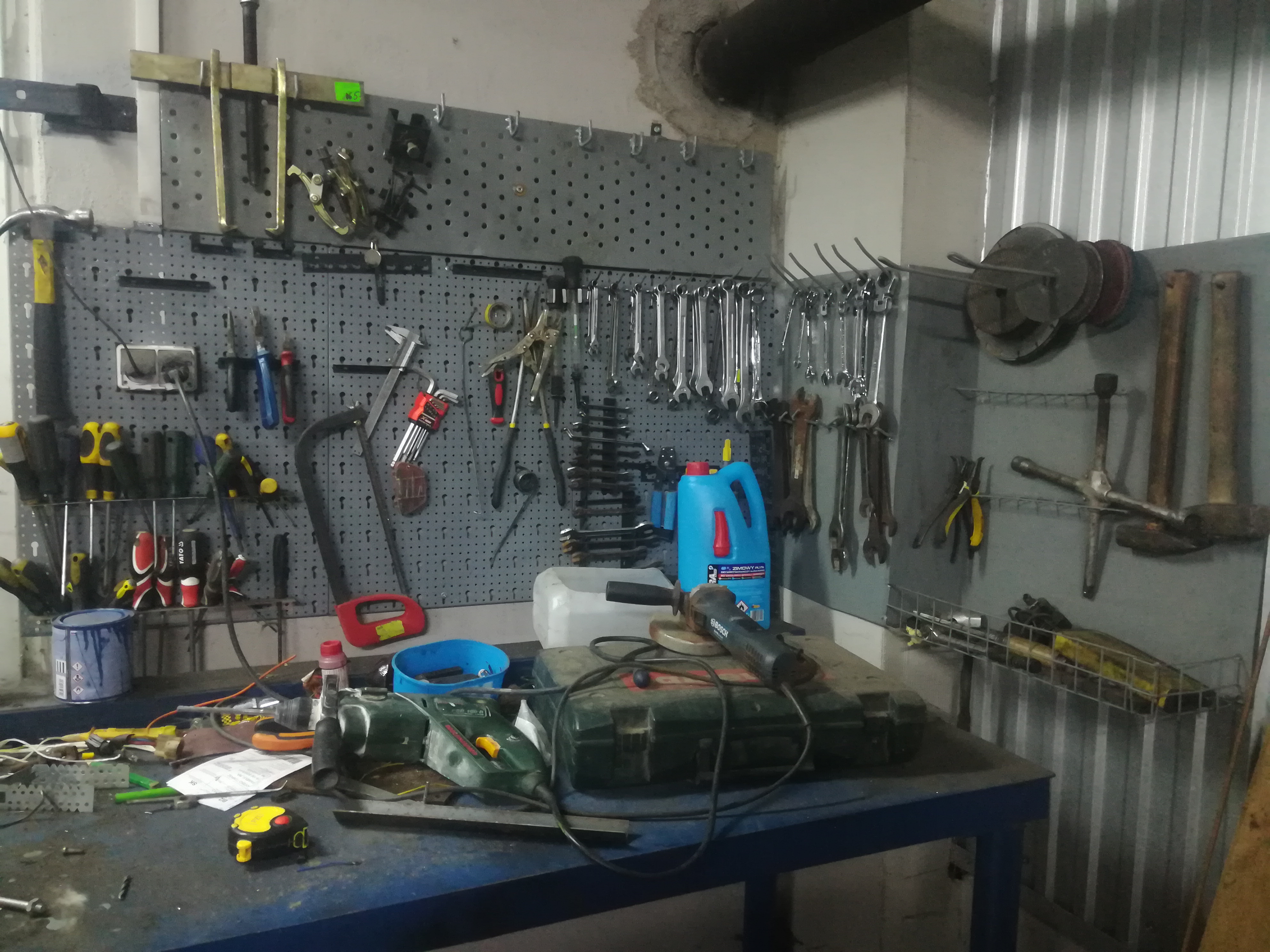 Warsztatowy stół roboczy z narzędziami, w tle ściana z perforowanej płyty z zawieszonymi kluczami, wkrętakami i innymi narzędziami, na stole widoczna szlifierka kątowa Bosch, wiertarka i miara...